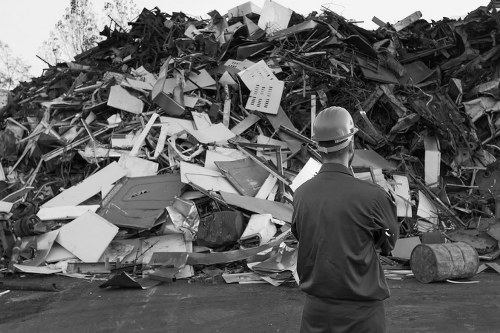 A man wearing a safety helmet and dark work clothing stands in front of a large pile of mixed construction and domestic waste, including broken furniture, wooden planks, metal scraps, and plastic items, all piled high on an outdoor surface, likely a driveway or yard. The waste collection appears disorganized with items haphazardly stacked, and the scene is captured during daylight, highlighting the textures and variety of materials in the debris. This image visually represents the type of rubbish that commercial waste removal services, such as those provided by Commercial Waste Removal Lewisham, assist businesses and property owners in disposing of responsibly. The setting appears to be an industrial or waste management area, emphasizing the importance of proper disposal practices for bulky, mixed waste materials commonly handled in Lewisham and surrounding areas. The environment suggests a focus on waste collection and clearance services aimed at clearing large quantities of rubbish efficiently and safely, consistent with professional disposal procedures in the local area.