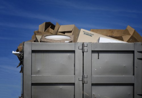 A large, open-top metal waste container filled with assorted cardboard boxes and packaging materials, situated outdoors against a clear blue sky. The cardboard boxes vary in size and are made of brown recycled paper, some flattened and others partly assembled, with visible printed text and logos on a few of them. The metal container is positioned on a paved surface, with its sides reinforced by horizontal and vertical panels, and appears to be part of a waste collection or rubbish removal service. The overall scene depicts a typical outdoor area used for rubbish disposal or waste collection, consistent with commercial waste removal services in Lewisham and nearby areas.