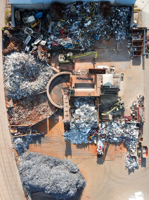 A view through a narrow gap between metal bars shows a cluttered outdoor area filled with discarded waste and scrap materials. In the foreground, the dark, textured bars frame the scene. Beyond, piled